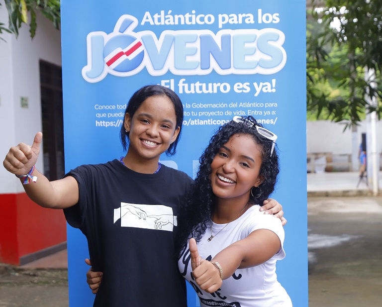 Dos mujeres jóvenes posando junto a la imagen del Programa Atlántico para los jóvenes.
